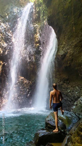 A man in swimsuit standing on the slippery rocks in front of Twin Waterfall  Bali  Indonesia. The waterfall is surrounded by rock from each side. The man enjoyins the beauty of the nature. Adventure