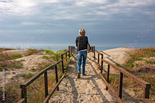 A tourist on the phone while walking on a boardwalk on the sand dunes, by the sea.