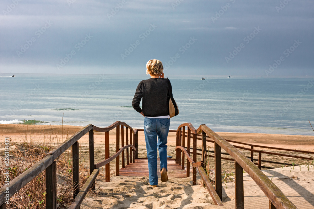 A tourist on the phone while walking on a boardwalk on the sand dunes, by the sea.