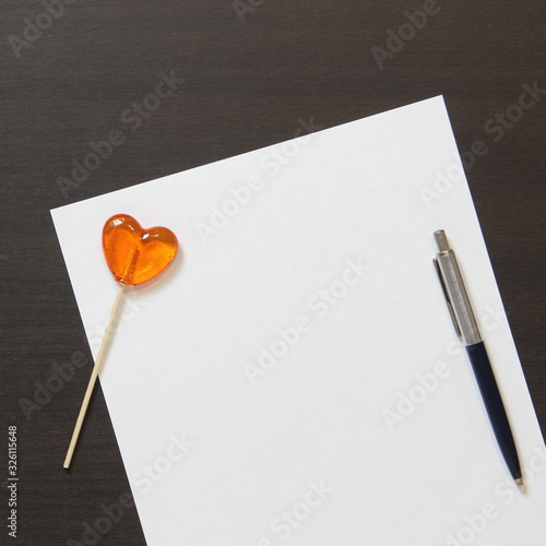 Template of white paper with pen and heart shaped caramel on dark wenge color wooden background.