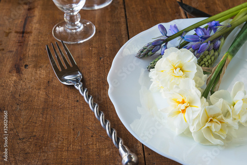 Festive table setting with forged fork and knife and bouquet of daffodils and hyacinths flowers on white plate. Vintage background, close up