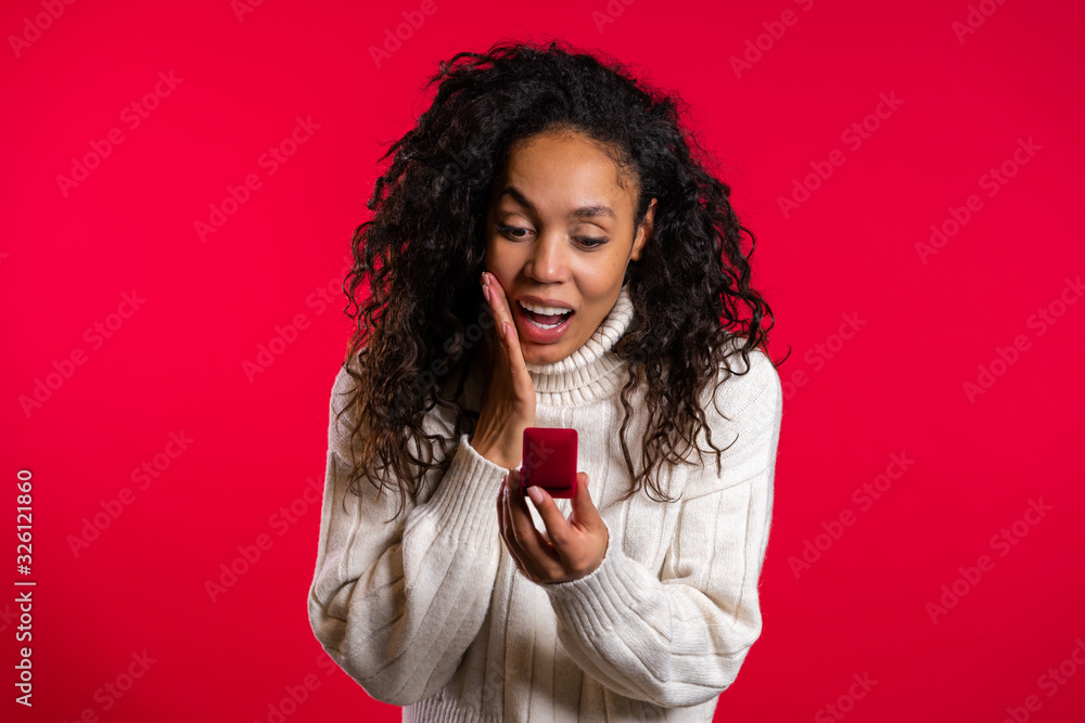 Naklejka premium Amazed african american young woman holding small jewelry box with proposal diamond ring on red wall background. Girl smiling, she is happy to get present, proposition for marriage.