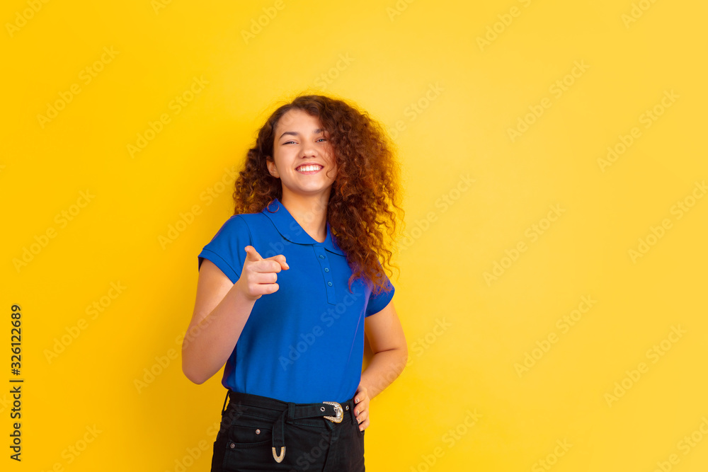 Pointing, laughting. Caucasian teen's girl portrait on yellow studio background. Beautiful female curly model in blue shirt. Concept of human emotions, facial expression, sales, ad. Copyspace. Pointing, laughting. Caucasian teen's girl portrait on yellow studio background. Beautiful female curly model in blue shirt. Concept of human emotions, facial expression, sales, ad. Copyspace.