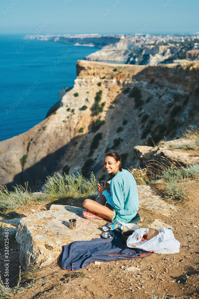 Full length portrait of carefree smiling woman sitting on cliff edge, having a breakfast outdoor on mountain peak with beautiful sea view.