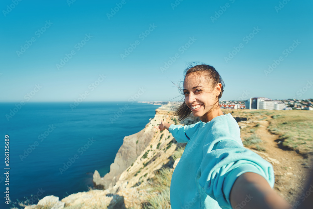 young female tourist making selfie and smile to the camera on cliff edge with beautiful sea view and enjoying wonderful nature.