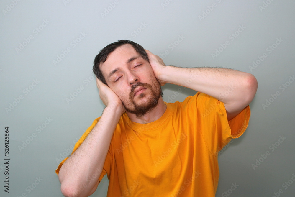 Middle-aged man with a mustache and a brunette beard in a yellow T-shirt on a gray background with closed eyes he covered his ears with the palms of his hands and bent as if he was listening to music Middle-aged man with a mustache and a brunette beard in a yellow T-shirt on a gray background with closed eyes he covered his ears with the palms of his hands and bent as if he was listening to music