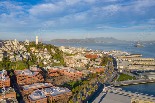 San Francisco, California, daytime, sunrise, aerial view of North Beach with Coit Tower and Golden Gate Bridge visible. Blue sky with clouds and golden light. Embarcadero area in foreground. 