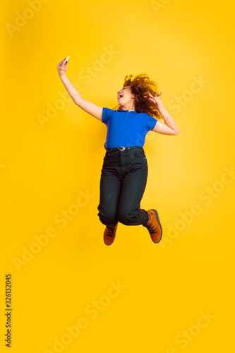 Jumping high, taking selfie. Caucasian teen's girl portrait on yellow studio background. Beautiful female curly model. Concept of human emotions, facial expression, sales, ad, education. Copyspace.
