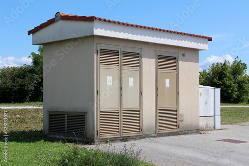 Side view of medium size local electricity substation building with metal front doors next to small grey plastic electrical box next to paved parking lot surrounded with grass and trees in background