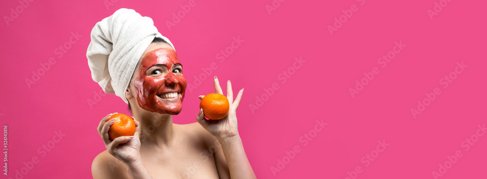 Fototapeta premium Beauty portrait of woman in white towel on head with red nourishing mask on face. Skincare cleansing eco organic cosmetic spa relax concept. A girl stands with her back holding an orange mandarin.