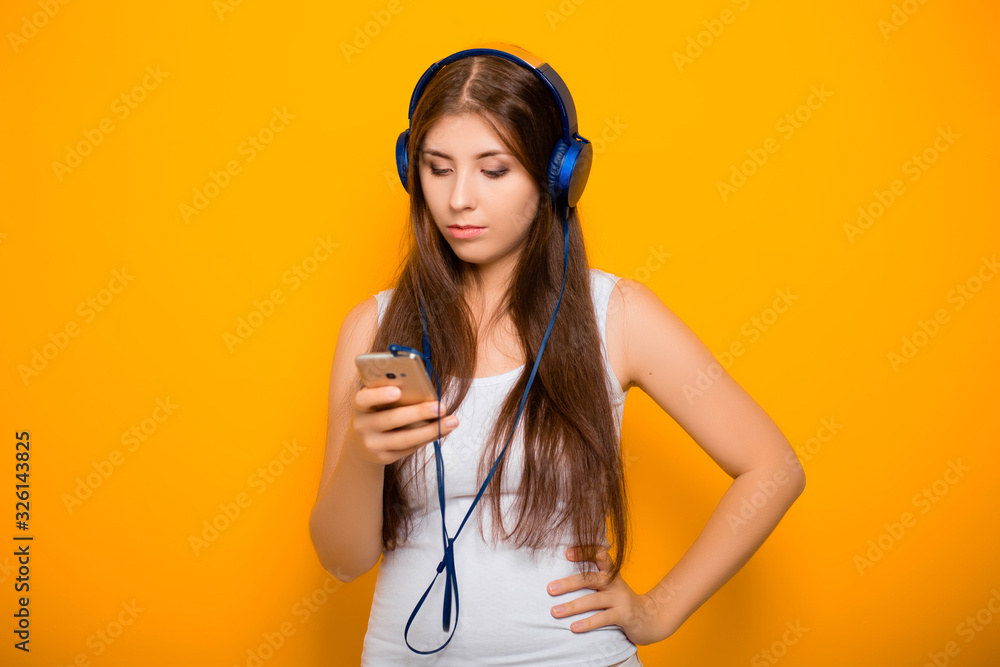 Attractive, young woman with a smartphone and in headphones, listens to music while standing on a yellow background. Attractive, young woman with a smartphone and in headphones, listens to music while standing on a yellow background.
