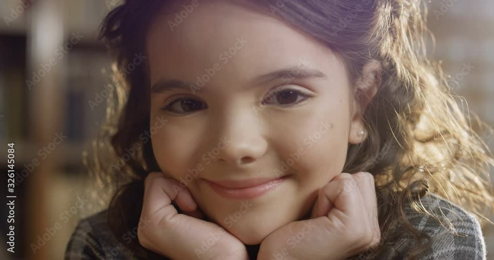 Portrait shot of Caucasian cute small girl with curly brunette hair smiling cheerfully to camera and leaning chin on hands in library. Close up of joyful little happy kid in bibliotheca. Portrait shot of Caucasian cute small girl with curly brunette hair smiling cheerfully to camera and leaning chin on hands in library. Close up of joyful little happy kid in bibliotheca.