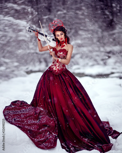 Amazing cute slim young woman in chic fairy tale image in red puffy dress and with crown on her head holds beautiful white violin and bow against background of white snow covered forest