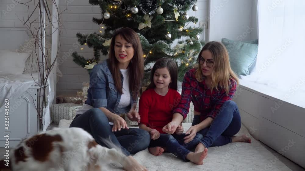 Two women and little girl with dog hugging near Christmas tree Two women and little girl with dog hugging near Christmas tree
