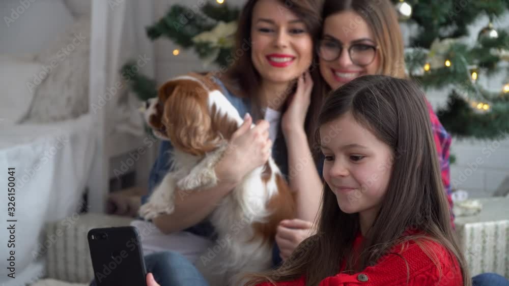 Two women and little girl with dog take a selfie near Christmas tree Two women and little girl with dog take a selfie near Christmas tree