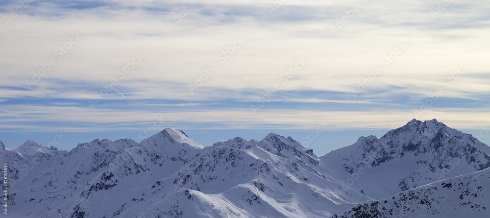 Fototapeta premium Snowy slope in high winter mountains and sunlit cloudy sky at evening