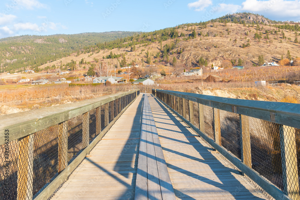 Looking across wooden trestle bridge on Kettle Valley Rail Trail to view of vineyards and mountains in autumn