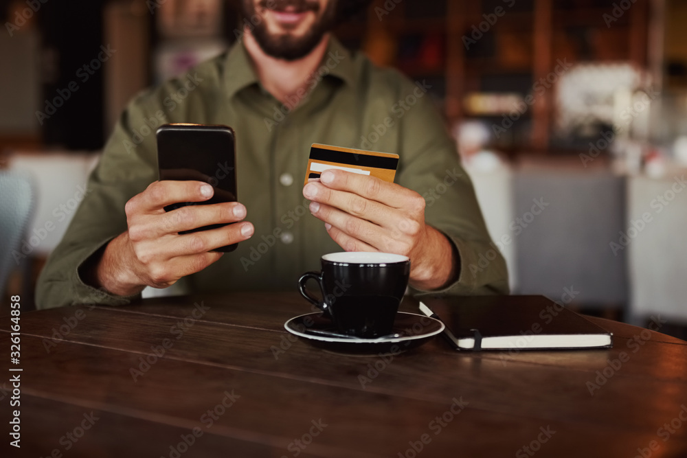 Closeup of cheerful happy caucasian man hand holding mobile phone typing card details to make online payment sitting in cafe