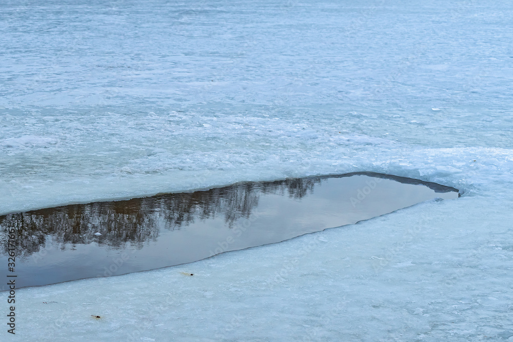 An ice hole in a frozen pond begins to melt. Trees and sky are reflected in the water. Spring is coming. Russia