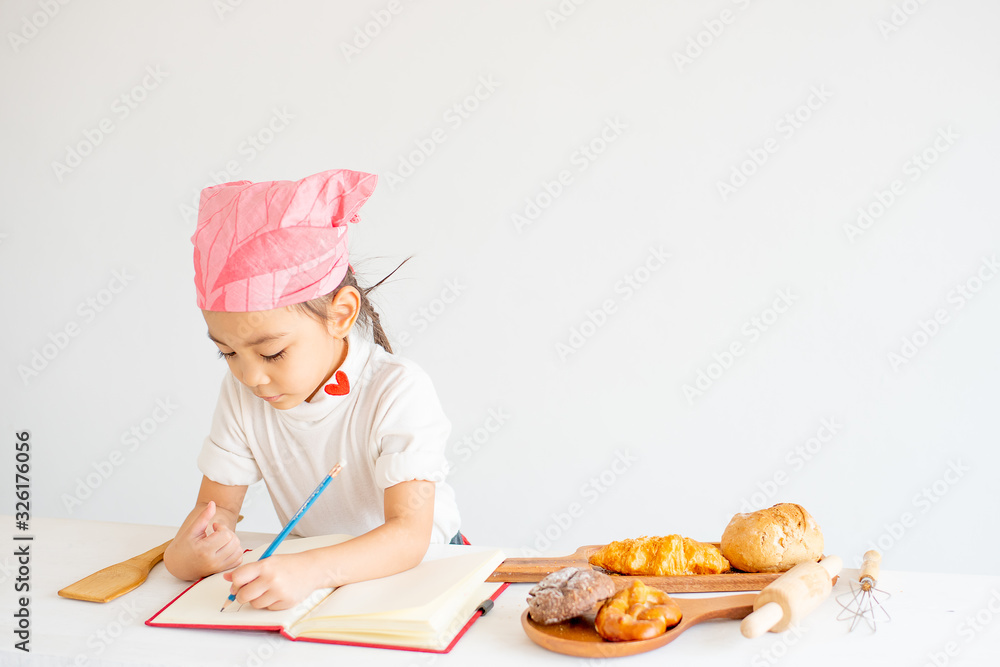 Asian little chef girl prepare and record her recipes about bread and cake cooking on notebook with white background. Asian little chef girl prepare and record her recipes about bread and cake cooking on notebook with white background.
