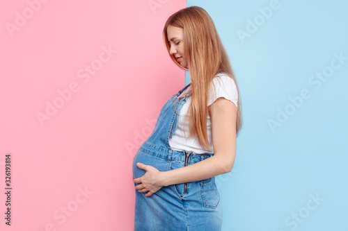 Happy young pregnant girl strokes her belly with her hands while standing on a pink and blue background