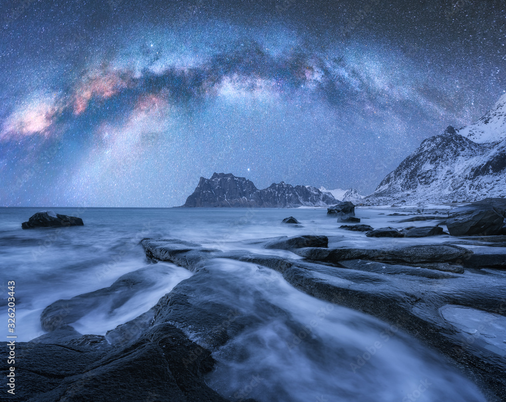 Milky Way over the snow covered mountains and rocky beach in winter at night in Lofoten Islands, Norway. Landscape with blue starry sky, water, stones, snowy rocks, bright milky way. Beautiful space