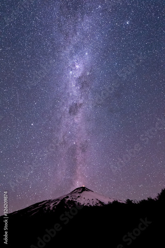 Thousands of stars in the sky with the Milky Way above the Villarrica Volcano in Chile with silhouettes of some trees, vertical photo