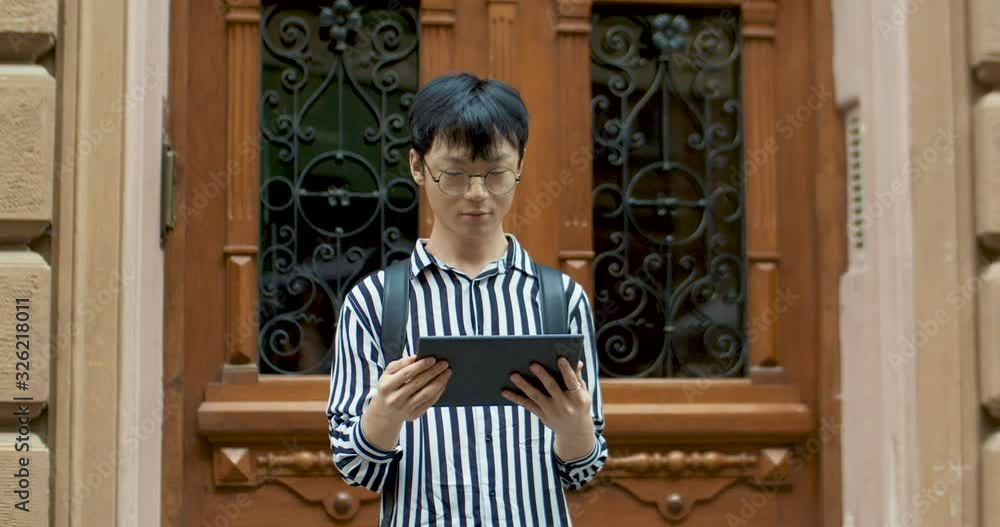 Happy Asian young male student in glasses and with bag over back standing outdoor at wooden door and watching video on tablet device. Chinese joyful man with computer in hands. Happy Asian young male student in glasses and with bag over back standing outdoor at wooden door and watching video on tablet device. Chinese joyful man with computer in hands.