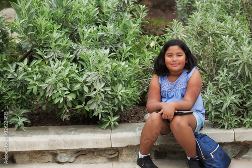 Tablou pe pânză An elementary school girl who is sitting down while she waits with her tablet and school backpack, surrounded by green plants