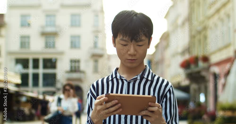 Chinese young man tapping and playing game on tablet device while standing at street in center of european city. Asian guy student typing and looking at map on computer outdoors. Traveling concept. Chinese young man tapping and playing game on tablet device while standing at street in center of european city. Asian guy student typing and looking at map on computer outdoors. Traveling concept.