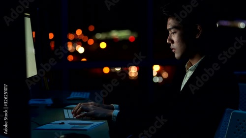 4K Overworked and stressed young Asian business man office worker working late alone at night in modern office. Tired businessman using computer working overtime for business project deadlines. photo
