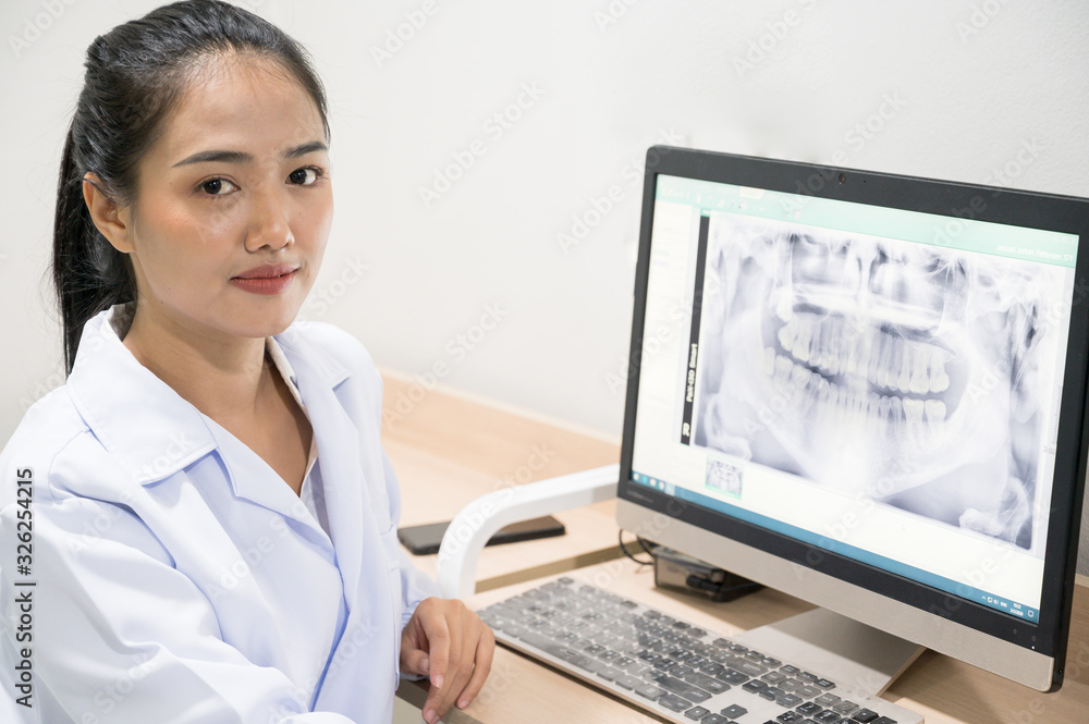 Portrait Asian woman dentist sitting and looking tooth image x-ray in computer.Female doctor examining patient tooth with panoramic radiograph on dental chair at the dental office. Portrait Asian woman dentist sitting and looking tooth image x-ray in computer.Female doctor examining patient tooth with panoramic radiograph on dental chair at the dental office.