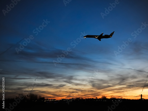 Silhouette of passenger plane flying over dark field with soft glow of city lights, wispy clouds and orange red sunset in background.