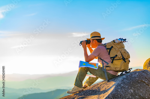 The young man with a lifted backpack looked in binoculars sitting on top of the mountain, enjoying a beautiful view of the mountains above the clouds.