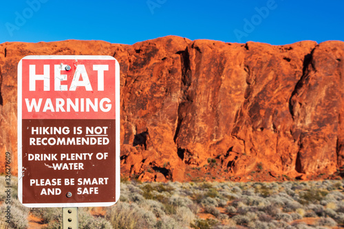 Heat warning sign informs travelers of the dangers of heat exhaustion from hiking in the desert climate. Blurred scenic red sandstone formations and desert scrub landscape background. Blue sky photo