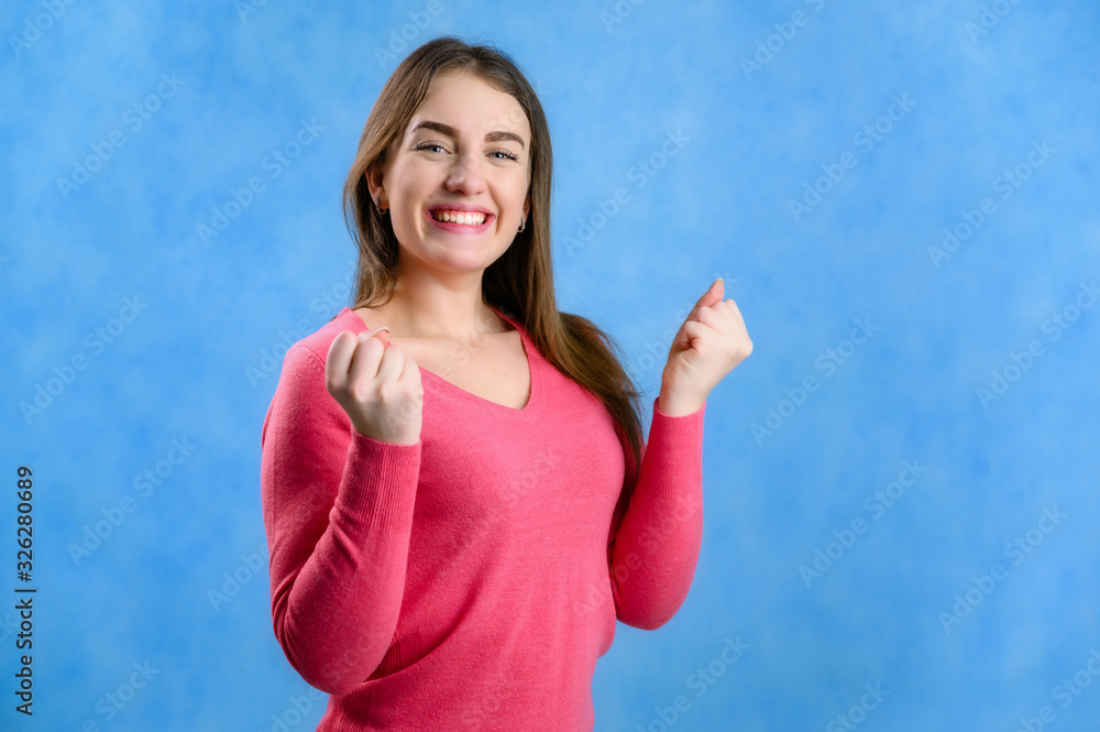 Fototapeta premium Portrait on a blue background of a pretty brunette Caucasian woman in a red sweater shows joy and happiness with a smile
