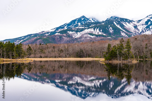 Small islet in the lake, beautiful lake surface reflecting blue sky like a mirror, rolling mountain range and woodland in the background on springtime sunny day. High latitude country natural scenery