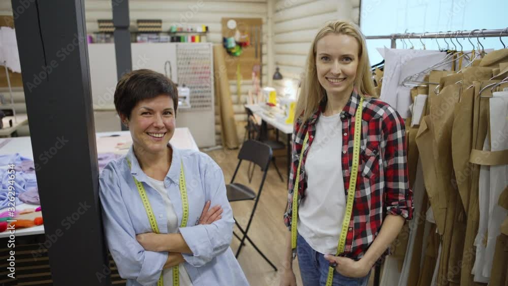 Tracking medium shot portrait of two female tailors, mother and adult daughter, looking at camera and smiling while posing near rail with sewing patterns in dressmaking studio Tracking medium shot portrait of two female tailors, mother and adult daughter, looking at camera and smiling while posing near rail with sewing patterns in dressmaking studio