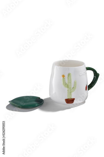 Subject shot of a white ceramic mug with a picture of a flowered cactus in a pot and a ladybird. The cute cup with a figured handle and a green leaf-formed cover are isolated on the white background. 