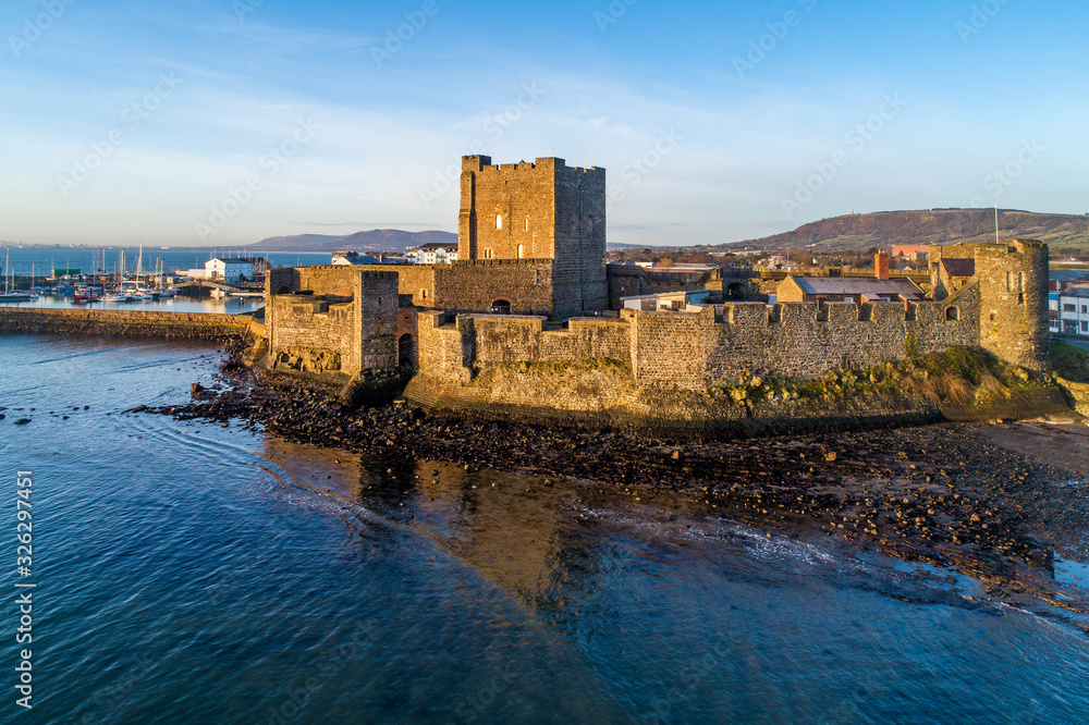 Medieval Norman Castle in Carrickfergus near Belfast in sunrise light. Aerial view with marina, yachts,  breakwater, groyne, sediments and far view of Belfast in the background
