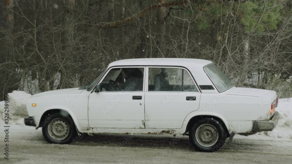 an old dirty white car on the background of a gray forest in winter in a snowfall an old dirty white car on the background of a gray forest in winter in a snowfall