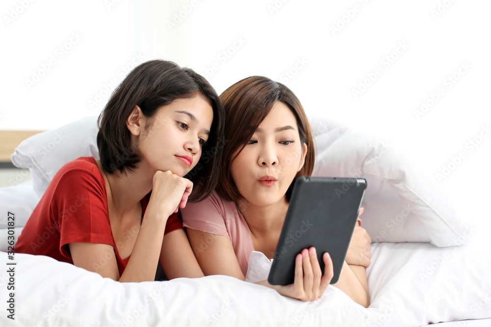 Fototapeta premium Happy young couple friend beautiful asia women lying on the bed in a bedroom and using digital tablet.