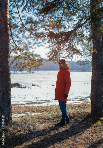 A young man in a red jacket on the background of the lake. Pine forest.