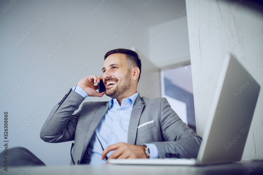 Cheerful handsome classy caucasian businessman in suit holding in one hand eyeglasses and using smart phone for business call. On table is laptop. Cheerful handsome classy caucasian businessman in suit holding in one hand eyeglasses and using smart phone for business call. On table is laptop.