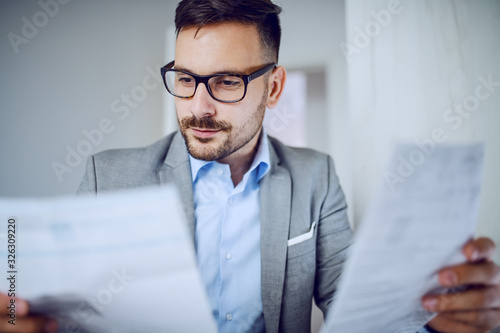 Head shot of hardworking sophisticated caucasian businessman in suit and with eyeglasses looking at documents while sitting in his office.