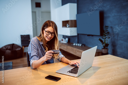 Young attractive smiling Caucasian woman with eyeglasses holding credit card and typing number of her bank account on laptop while sitting at dining table. Online shopping concept.