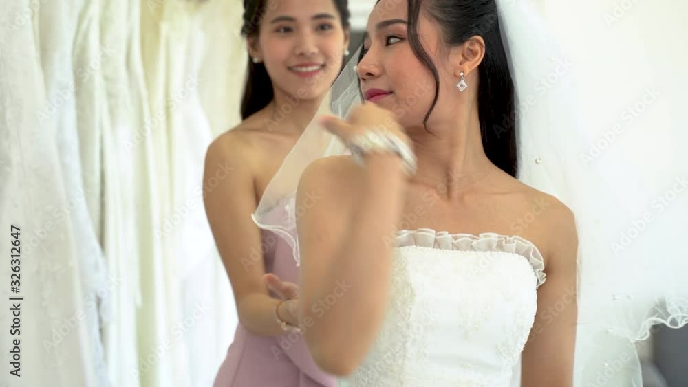 Pretty girl is trying a beautiful wedding dress on and looking at the mirror in a wedding salon. Friend is helping her with a choice. Mirror has a Christmas theme ornamentation. Pretty girl is trying a beautiful wedding dress on and looking at the mirror in a wedding salon. Friend is helping her with a choice. Mirror has a Christmas theme ornamentation.