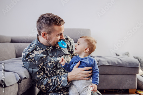Attractive bearded soldier sitting on the floor in living room, holding his beloved adorable baby boy, holding teether in his mouth and teasing him.