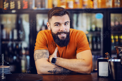 Foto Portrait of handsome bearded smiling positive tattooed barman leaning on bar counter, looking at camera and waiting for customers to order drinks
