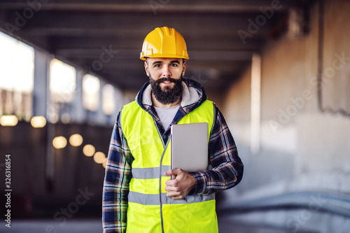 Εκτύπωση καμβά Portrait of highly motivated caucasian hardworking smiling bearded supervisor with helmet on head in vest and with laptop in hands posing on construction site